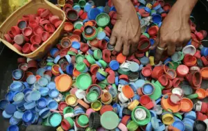 Various colored plastic lids spread out on the ground, with two hands sorting through them, suggesting an activity related to recycling or repurposing. Plastic Pollution Management